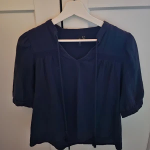 Mörkblå blus från Vero Moda - Säljer en stilren mörkblå blus från Vero Moda. Blusen har korta ärmar och en v-ringad design med knytband vid halsen. Perfekt för en elegant och avslappnad look.