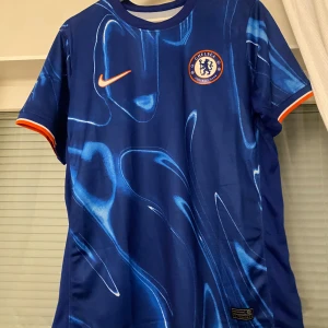 Blå Chelsea fotbollströja - Snygg blå Chelsea fotbollströja från Nike med ett unikt mönster i olika blå nyanser. Tröjan har korta ärmar med orange detaljer vid ärmsluten och klubbens emblem på bröstet. Perfekt för alla Chelsea-fans! Helt ny