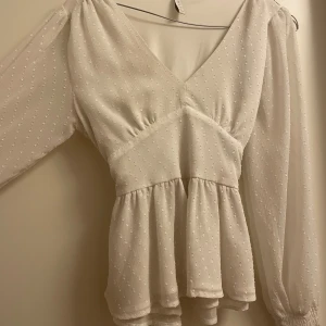 Vit blus från Amisu - Säljer en söt vit blus från Amisu med v-ringning och knytband i ryggen. Blusen har långa ärmar med elastiska muddar och ett diskret prickigt mönster. Perfekt för en stilren look.