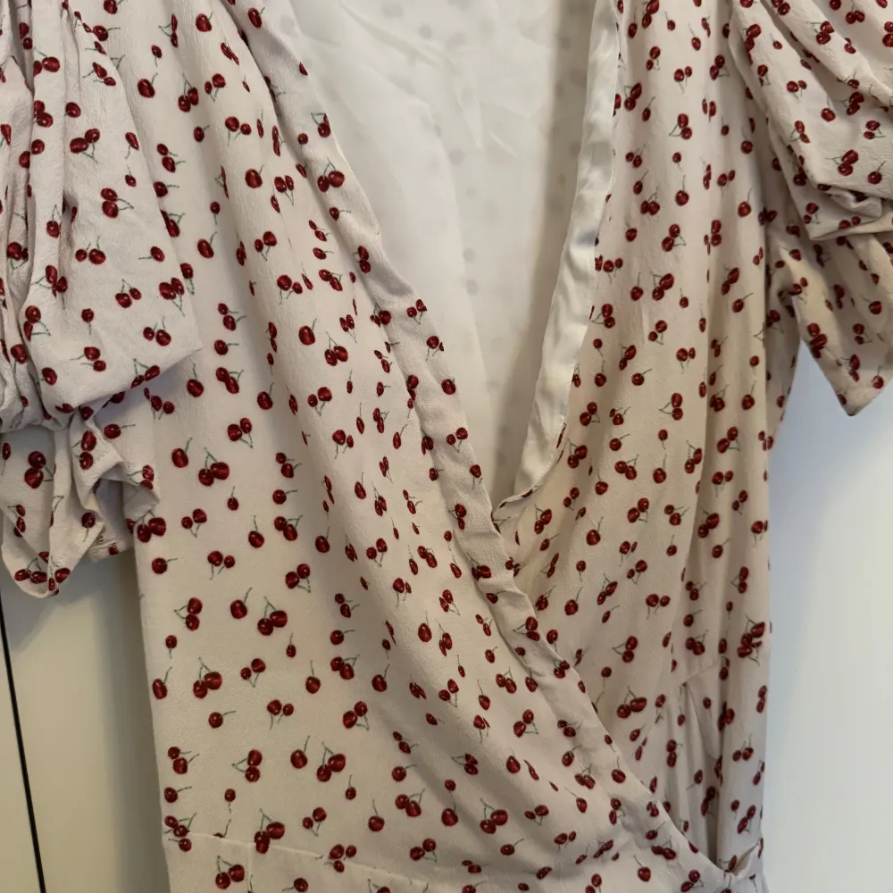 Charmig vit långklänning med röda körsbärsmönster. Klänningen har korta ärmar och en omlott-design med knytband i midjan. Använd en gång. Storlek 38.. Mekot.