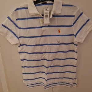 Oanvänd/ny Vit och blå randig ralph lauren pikétröja - Säljer en klassisk vit pikétröja från Ralph Lauren med blå ränder och den ikoniska loggan broderad på bröstet. Tröjan har korta ärmar och en krage med knappar. Perfekt för en stilren och avslappnad look.