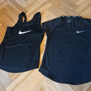 Svarta träningstoppar från Nike - Två svarta träningstoppar från Nike. Ena är en ärmlös linnetopp med brottarrygg och det klassiska Nike-logotypen på bröstet. Den andra är en kortärmad topp med v-ringning och samma loggtyp. Båda är i mycket bra skick