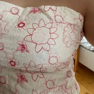 Vit tubtopp med blommigt mönster - Säljer en söt vit tubtopp med broderade blommor i rött och rosa. Perfekt för sommardagar och festivaler. Toppen är axelbandslös och har ett unikt blommigt mönster som ger en bohemisk känsla.