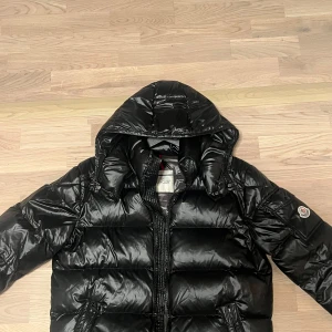 Svart pufferjacka från Moncler - Snygg svart pufferjacka från Moncler med glansig yta och dragkedja framtill. Jackan har en quiltad design och långärmade ärmar. Perfekt för kyliga dagar med sin varma och stilrena look.