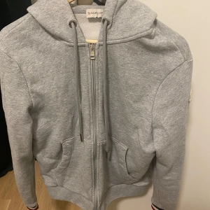 Grå Moncler kofta - Riktigt snygg moncler kofta som har långa ärmar och ribbade muddar med röd och blå randdetalj. Säljer då jag inte använder längre. Helt ren och i perfekt skick. Koftan är dubbelzip kommer även moncler påse med köpet, Finns mer bilder. Pris kan diskuteras 