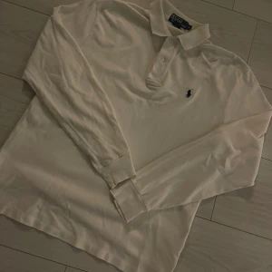 Vit pikétröja från Polo Ralph Lauren (S) - Klassisk vit pikétröja från Polo Ralph Lauren med den ikoniska loggan broderad på bröstet. Tröjan har långa ärmar och en stilren krage med knappar. Perfekt för en avslappnad men ändå stilfull look.