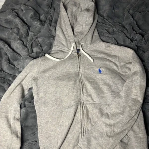 Grå hoodie från Ralph Lauren - Säljer en stilren grå hoodie från Ralph Lauren med dragkedja och huva. Den har en liten blå logga på bröstet och vita snören vid huvan. Perfekt för en avslappnad look. Dom går för va 3000. Har använt den några gånger, byt stil så det passar inte mig längre 
