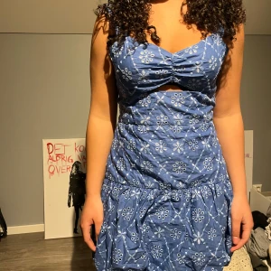 Blå blommig klänning - Charmig blå klänning med blommigt mönster och volangdetaljer. Klänningen har en v-ringad design med knyt framtill och är ärmlös. Perfekt för sommarens varma dagar.