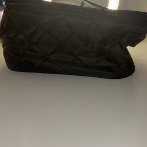 Svart quiltad necessär - Snygg och praktisk svart quiltad necessär med dragkedja. Insidan är ljus och har flera fack för organisering. Perfekt för att hålla ordning på dina saker när du är på resande fot.finns en liten fläck i en av insidorna.