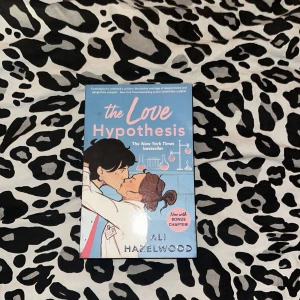 The Love Hypothesis - En populär roman av Ali Hazelwood, 'The Love Hypothesis', med en charmig illustration på omslaget. Boken handlar om en fejkad relation mellan forskare och utforskar teman som attraktion och vetenskap. Innehåller en bonuskapitel.