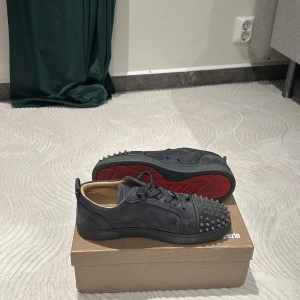 Gråa sneakers med nitar från Christian Louboutin - Snygga gråa sneakers från Christian Louboutin med nitar på ovansidan och den ikoniska röda sulan. Har haft skorna i en vecka och säljer dom nu för att ha spelar mycket fotboll och skorna förstörs tyvärr så på grund av de så är en nitt tyvärr borta men är fortfarande i jätte bra skick .Skorna har snörning och är tillverkade i skinn, vilket ger en lyxig känsla. Perfekta för att ge din stil en edgy touch.