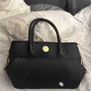 Svart handväska med gulddetaljer - Snygg svart handväska med gulddetaljer och dragkedja. Perfekt för att bära dina dagliga nödvändigheter med stil. Den har två handtag och en elegant design.