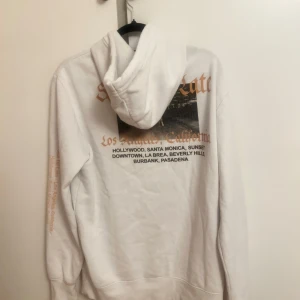 Vit hoodie med tryck - Säljer en vit hoodie med tryck på både fram- och baksidan. Hoodien har en stor ficka framtill och text i orange och svart. Perfekt för en avslappnad stil.