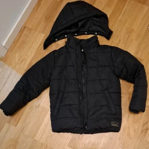 Svart pufferjacka från KappAhl - Säljer en svart pufferjacka från KappAhl med avtagbar huva och dragkedja framtill. Jackan har långa ärmar och är perfekt för kyligare väder. Den quiltade designen ger extra värme och stil. Litet, litet hål på ena ärmen som knappt syns och som lätt kan sys igen