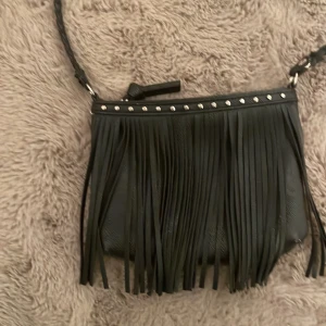 Svart axelväska med fransar - Snygg svart axelväska med fransar och nitar upptill. Perfekt för att ge din outfit en bohemisk touch. Väskan har en lång axelrem och är tillverkad i syntetmaterial. 40kr + frakt 