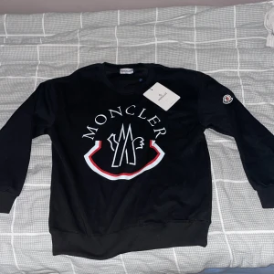 Svart sweatshirt från Moncler - Snygg svart sweatshirt från Moncler med stort tryck på framsidan. Tröjan har långa ärmar och en rund halsringning. Perfekt för en stilren och avslappnad look.
