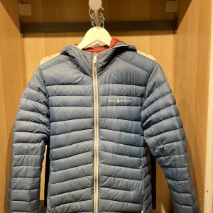 Blå pufferjacka från Sail Racing - Snygg blå pufferjacka från Sail Racing med dragkedja och huva. Jackan har en quiltad design och är perfekt för kyligare väder. Den har en logotyp på bröstet och en på ärmen.