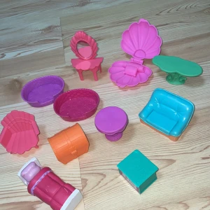 Färgglada plastmöbler för dockhus - Säljer ett set med färgglada plastmöbler för dockhus. Inkluderar en turkos soffa, rosa sminkbord med spegel, grön bord, lila bord, och fler tillbehör som en orange skattkista och rosa snäckformad stol. Perfekt för att inreda ett fantasifullt dockhus!