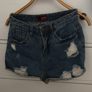 Blå jeansshorts från My Style - Snygga blå jeansshorts med slitna detaljer från My Style. De har en klassisk femficksdesign och knappgylf. Perfekta för en avslappnad look.
