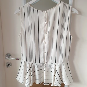 Vit och svart randig blus från Zara - Snygg ärmlös blus från Zara i vitt med svarta ränder. Blusen har en peplumdetalj i midjan och knappar på baksidan. Perfekt för en stilren look.
