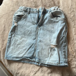 Ljusblå jeanskjol  - Trendig ljusblå jeanskjol med slitningar framtill. Kjolen har en klassisk femficksdesign och en bekväm resår i midjan. Perfekt för en avslappnad stil.