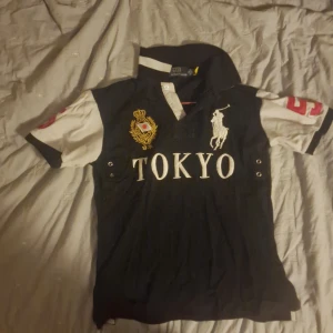 Svart och vit pikétröja från Polo Ralph Lauren - Priset kan variera. Svår att hitta TOKYO Ralph Lauren Pike/polo. Snygg svart pikétröja från Polo Ralph Lauren med vita ärmar och stora röda siffror. Tröjan har texten 'TOKYO' på framsidan och en broderad logga. Perfekt för en sportig look!