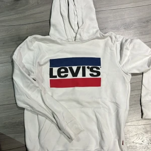 Vit hoodie från Levi's - Säljer en vit hoodie från Levi's med det klassiska logotyptrycket i blått och rött på framsidan. Tröjan har en bekväm passform med långa ärmar och en praktisk ficka framtill. Knappt använd!