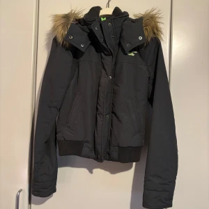 Grå vinterjacka från Hollister - ! Säljer en grå vinterjacka från Hollister med pälskantad huva och grönt foder. Jackan har dragkedja och knappar framtill samt ribbade muddar vid ärmslut. Köpt på holloster runt 2011-12