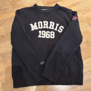 Morris tröja - Säljer en svart tröja från Morris med texten 'Morris 1968' i vitt på framsidan. Tröjan har en v-ringad design och ribbade muddar vid ärmslut och nederkant. Det finns en broderad detalj med en flagga på ärmen.