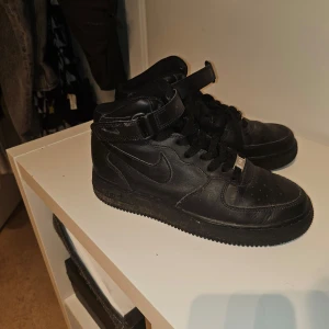 Svarta Nike Air Force med snörning - Säljer ett par klassiska svarta Nike Air Force med hög modell och snörning. Skorna har en stilren design med en justerbar rem vid ankeln för extra stöd. Perfekta för en sportig look. Behöver endast lite rengöring!
