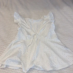 Vit top med volangdetaljer - Charmig vit top med volangdetaljer och knytning mitt på. Toppen har ett diskret hålmönster och korta ärmar. Perfekt för somriga dagar.
