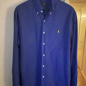 Blå skjorta från Ralph Lauren - Snygg blå skjorta från Ralph Lauren med klassisk krage och knappar framtill. Den har en broderad gul logotyp på bröstet och är i slim fit. Perfekt för en stilren look.