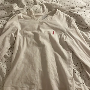 Vit långärmad tröja från Polo Ralph Lauren - Säljer en stilren vit långärmad tröja från Polo Ralph Lauren med det klassiska röda logotypbroderiet på bröstet. Perfekt för en avslappnad och snygg look. Tröjan är tillverkad i mjukt material för extra komfort.