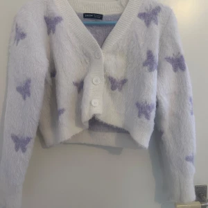 Vit och lila kofta från Shein - Mysig vit kofta från Shein med lila fjärilsmönster, i storlek S/36. Den har en croppad passform och knappar framtill. Perfekt för en stilren och bekväm look.