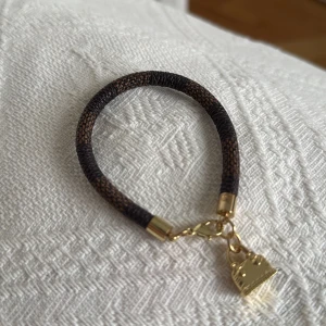 Brunt armband i klassiskt rutigt mönster med gulddetaljer - Snyggt brunt armband med klassiskt rutigt mönster (lv) gulddetaljer och en hängande guldfärgad väska. Perfekt accessoar för att ge en elegant touch till din outfit.