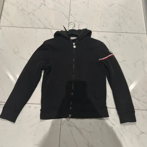 Svart huvtröja med dragkedja från Moncler - Snygg svart huvtröja från Moncler med dragkedja och broderad logga på ärmen. Tröjan har en stilren design med ribbade muddar och en röd-vit randdetalj på ärmen. Perfekt för en casual look.