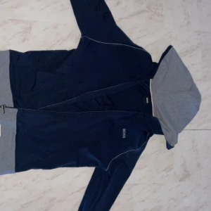 Mörkblå hoodie från Boss - Säljer en stilren mörkblå hoodie från Boss med grå insida på huvan. Den har dragkedja framtill och långa ärmar. Perfekt för en avslappnad look. Säljer på grund av att tröjan inte kommit till så mycket användning och är i mycket bra skick som ny nästan. Det är inga fläckar eller slitage på tröjan. Är det något ni undrar över är det bara att skriva. Ny pris mellan 750-900 ungefär 