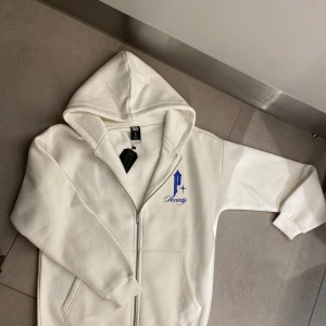 Vit hoodie från P-society - Snygg vit hoodie från P-society med blått tryck fram och bak. Framsidan har en stilren logga medan baksidan pryds av en stor duva och texten 'Paranoid'. Hoodien har dragkedja och är perfekt för en avslappnad stil. Har i alla storlekar, Skicka pm och välj din storlek! säljes för 400kr.