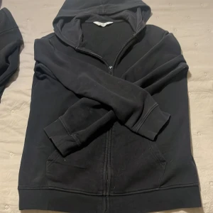 Svart hoodie från H&M - Säljer en klassisk svart hoodie från H&M med dragkedja och huva. Perfekt för en avslappnad stil. Tröjan har långa ärmar och är tillverkad i ett mjukt material för extra komfort.