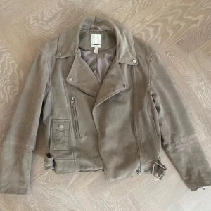 Beige mockajacka med dragkedja - Snygg beige mockajacka med dragkedja och bälte i midjan. Jackan har långa ärmar och en stilren design som passar perfekt för höst och vår. Perfekt för att ge din outfit en cool touch.