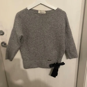 Trekvartsärmad stickad tröja från By o la la - Mysig grå tröja från By o la la med trekvartsärmar och en dekorativ svart rosett vid nederkanten. Perfekt för kyliga dagar och ger en stilren look. Storleken är S.