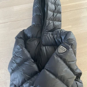 Svart pufferjacka från EA7 - Säljer en svart pufferjacka från EA7 med dragkedja och huva. Jackan har ett stilrent märke på ärmen och är perfekt för kyligare dagar. Den är långärmad och har en quiltad design som ger extra värme.