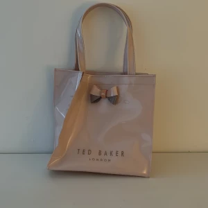 Ljus rosa shoppingväska från Ted Baker - Snygg ljus rosa shoppingväska från Ted Baker med en elegant rosett på framsidan. Väskan har två handtag och är perfekt för att bära med sig det nödvändigaste. Den blanka ytan ger en stilren look. På framsidan och baksidan ser man Ted Baker loggan i rosé guld