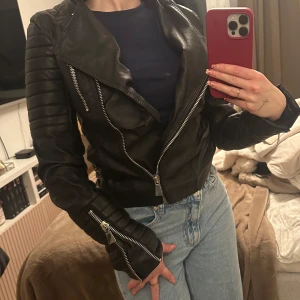 Svart skinnjacka med dragkedja - Snygg svart skinnjacka med coola dragkedjor och bältesdetaljer. Jackan har en tight passform och långa ärmar med dragkedjor vid ärmsluten. Perfekt för en edgy look! Storlek 36 men känns som XS/XSS då jag är petite och 160 cm lång, supr snygg tajt i midjan! Ger 2015 vibes hehe