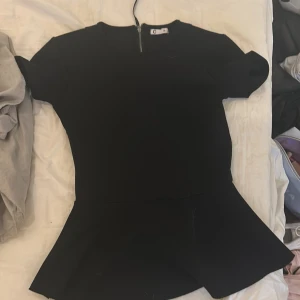 Svart peplumtopp - Snygg svart peplumtopp med dragkedja i ryggen. Den har korta ärmar och en figurnära passform som ger en stilren look. Perfekt för en kväll ute eller en speciell tillställning.