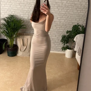 Beige långklänning med smala axelband - Elegant beige långklänning med smala axelband och en figurnära passform. Klänningen har en mjuk och flytande design som ger en stilren look. Perfekt för speciella tillfällen.
