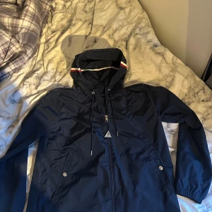 Mörkblå windbreaker från Moncler - Säljer en snygg mörkblå vindjacka från Moncler med luva och dragkedja. Jackan har en stilren design med Moncler-logga på ärmen och röd-vit randdetalj på huvan. Scan fungerar och den är äkta. Skriv vid minsta fundering. Perfekt för blåsiga dagar!