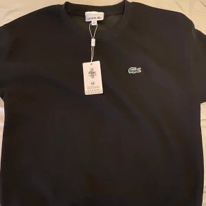 Svart sweatshirt från Lacoste - Säljer en stilren svart sweatshirt från Lacoste med det klassiska krokodilmärket på bröstet. Tröjan har rund hals och långa ärmar, perfekt för en avslappnad look. Passar bra till både jeans och chinos. Aldrig använd.