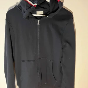 Svart hoodie med dragkedja från Moncler - Snygg svart hoodie från Moncler med dragkedja och randiga detaljer i rött och vitt på huvan. Perfekt för en stilren och bekväm look. Den har långa ärmar och en klassisk passform. Storlek m-L, 10/10 stick