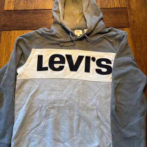 Grå hoodie från Levi's - Säljer en snygg grå hoodie från Levi's med stor logga i svart på bröstet. Tröjan har en justerbar huva med snören och en praktisk magficka. Använt denna i tre dagar